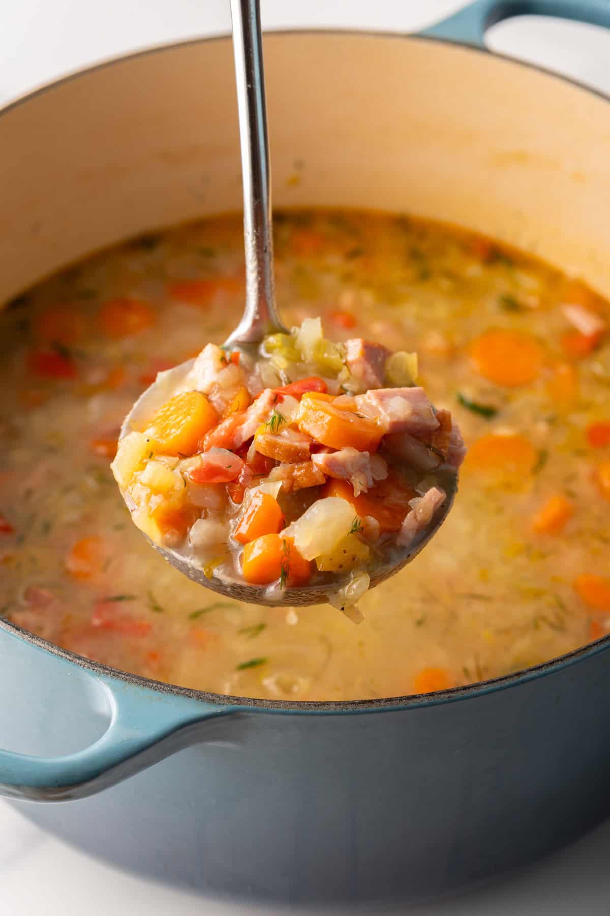 Metal ladle holding a scoopful of cabbage ham soup to camera, held over a blue cooking pot of soup.