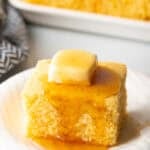 Square of cornbread is on a white plate, topped with a pat of butter and drizzled with honey.