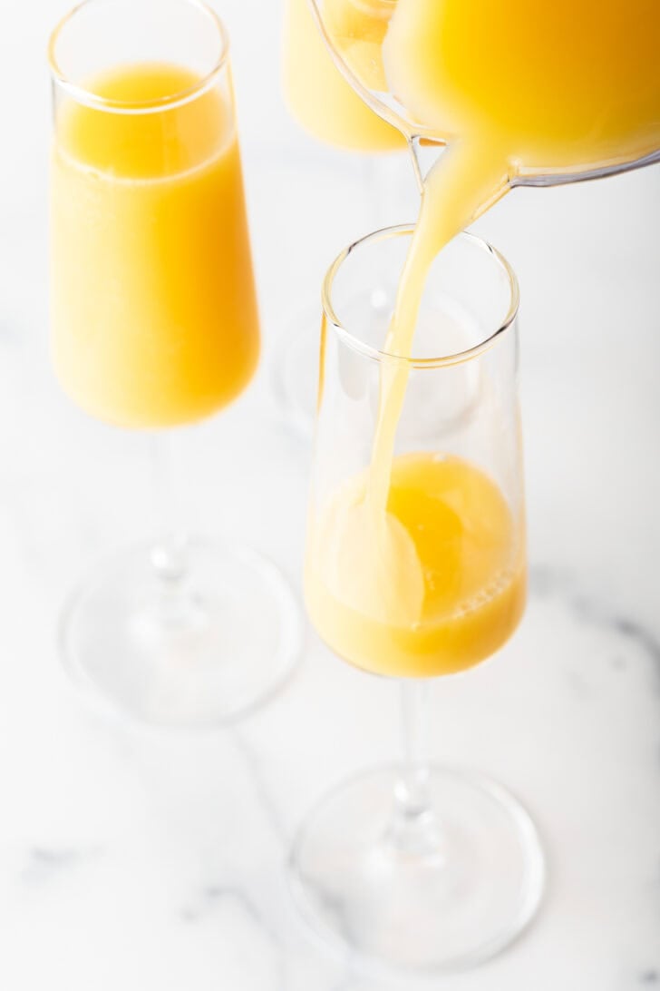 Pouring orange juice from a pitcher into a tall champagne glass.