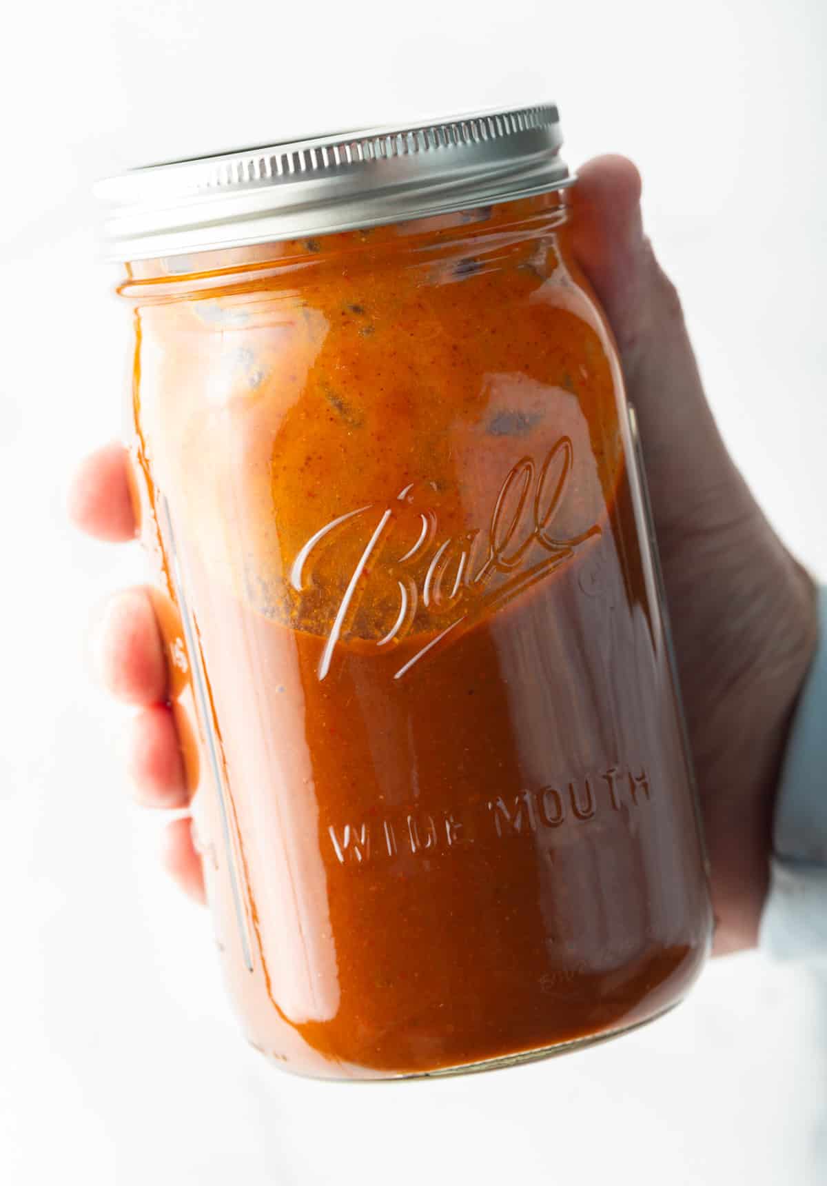 Hand holding a large glass mason jar filled with red enchilada sauce.