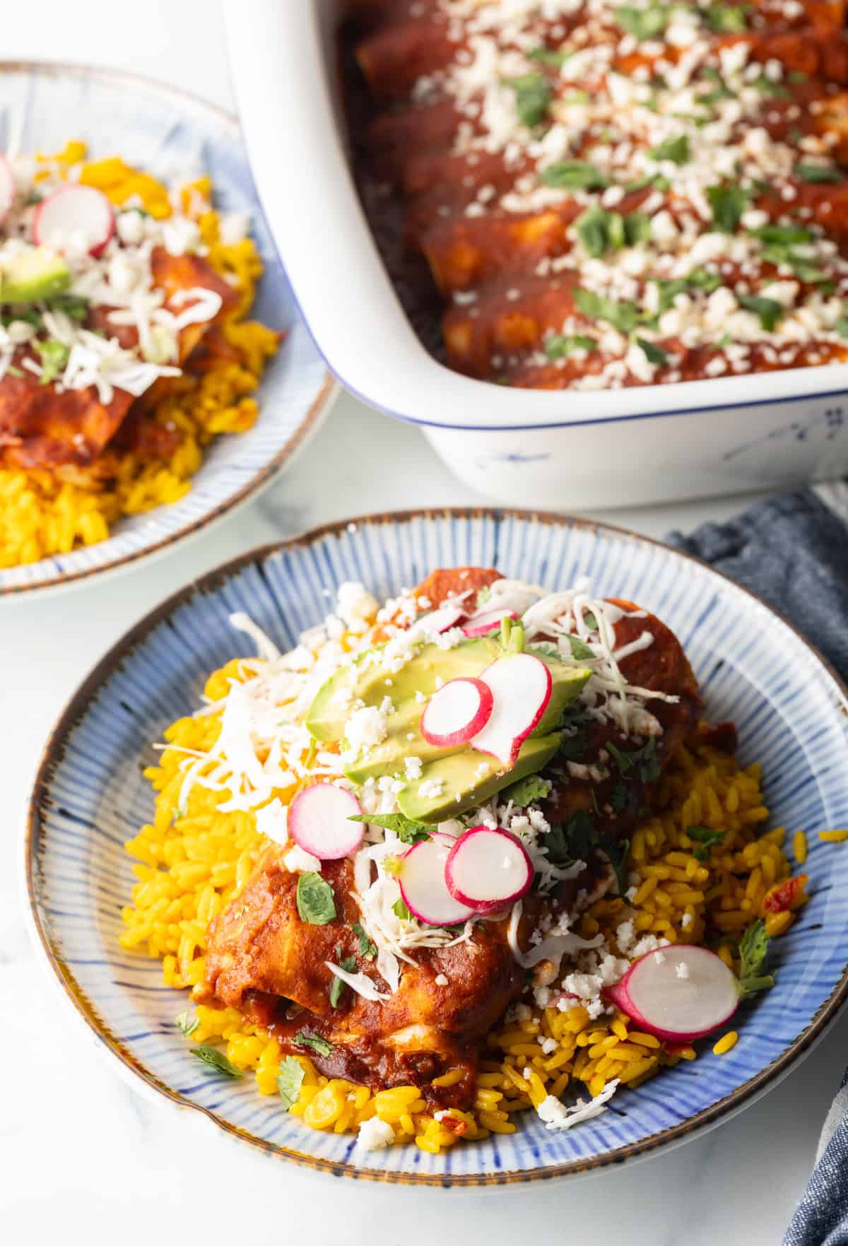 Large plateful of enchiladas with red sauce, topped with slices of radishes and fresh slaw, on a plate next to yellow rice. A large white baking dish of enchiladas is in the background.