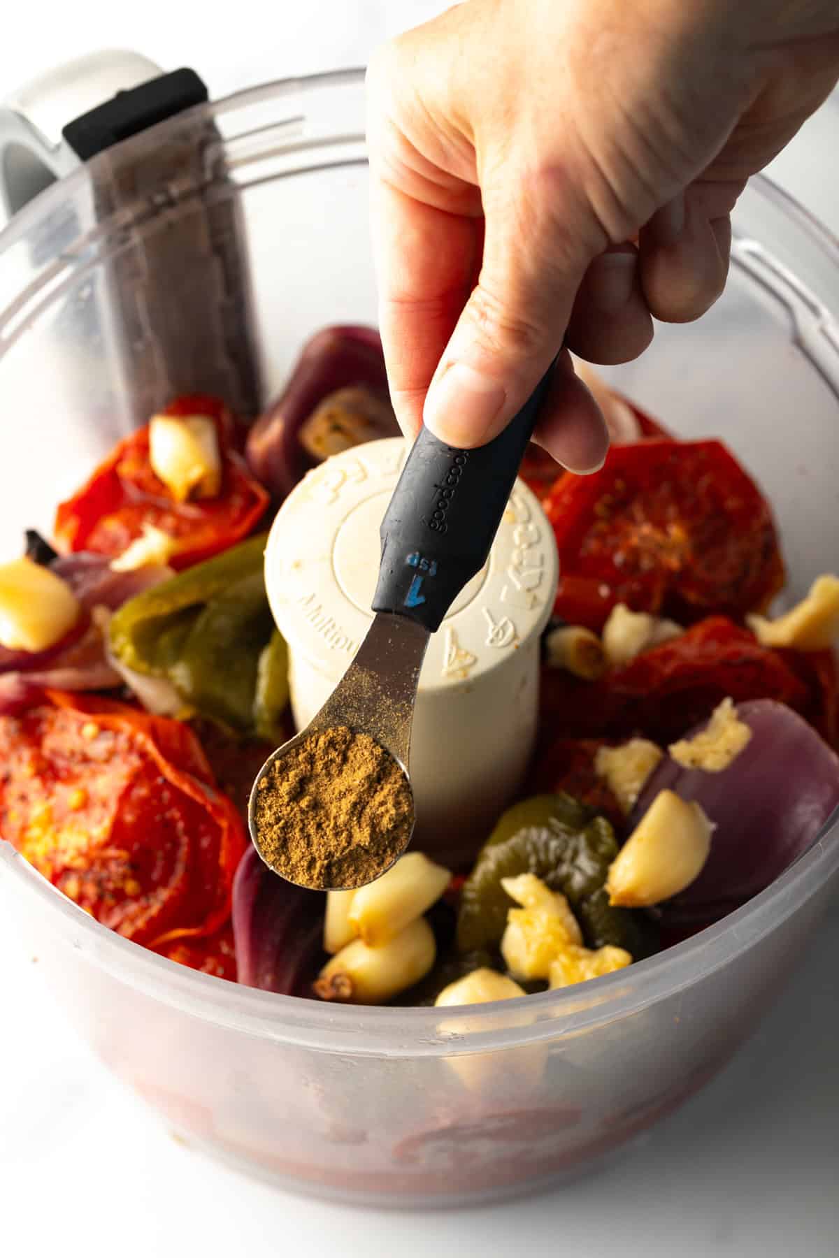Hand adding spices from a measuring spoon into the food processor with salsa ingredients.