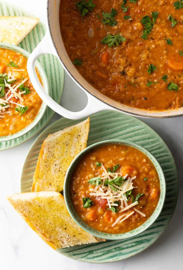 Top down view green bowl of carrot and red lentil soup topped with chopped herbs and shredded cheese. The bowl is on a plate with two baguette slices.