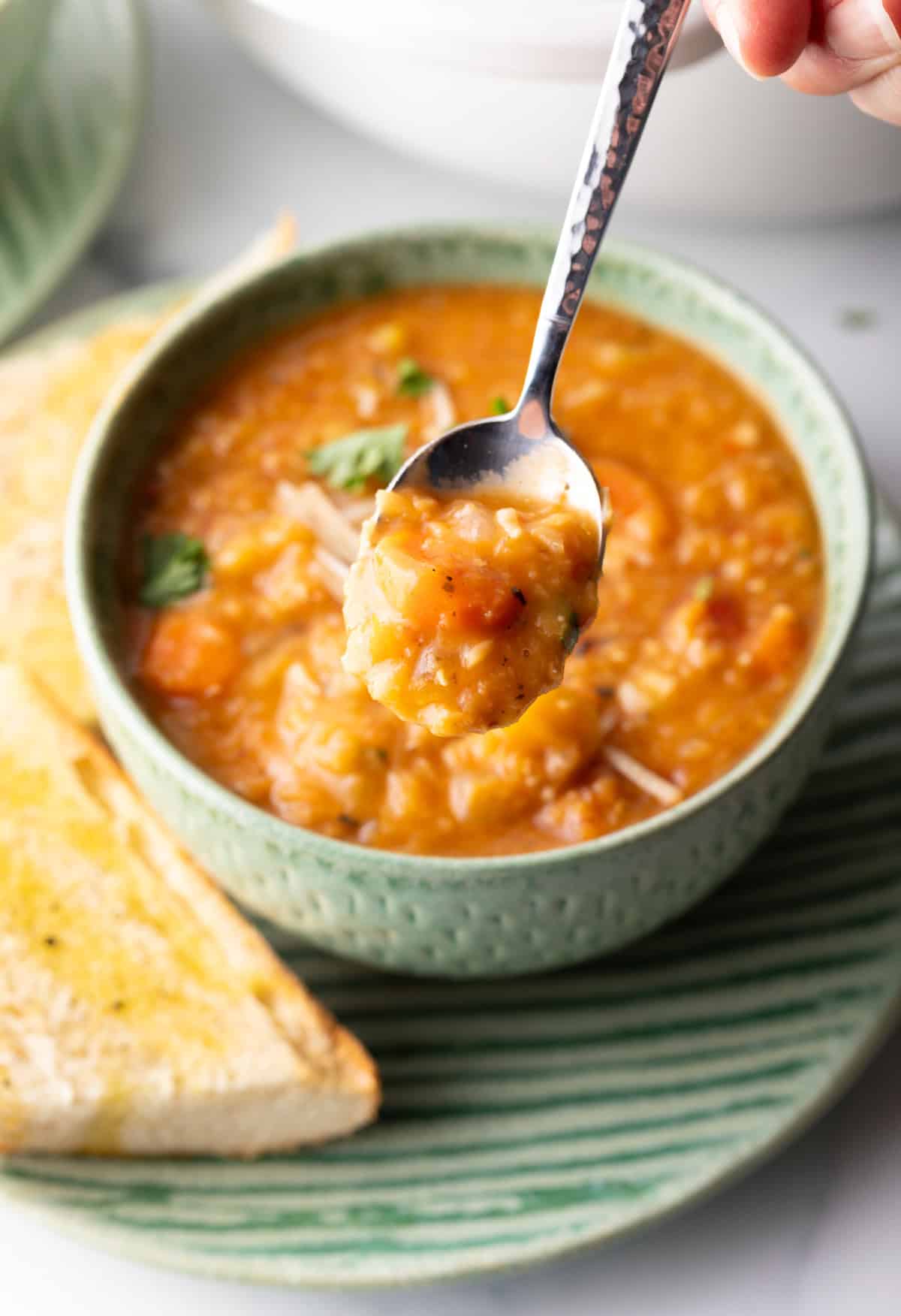 A hand is holding a spoonful of lentil soup to camera from a green bowlful.