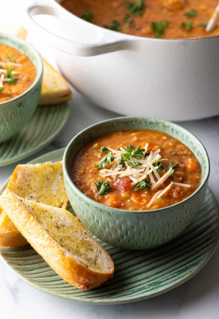 Green bowl of carrot red lentil soup topped with chopped herbs and shredded cheese. The bowl is on a plate with two baguette slices.