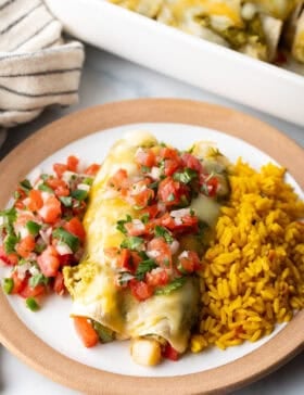White plate with a serving of veggie enchiladas with green sauce and pico de gallo, nestled between a serving of pico de gallo and spanish rice.