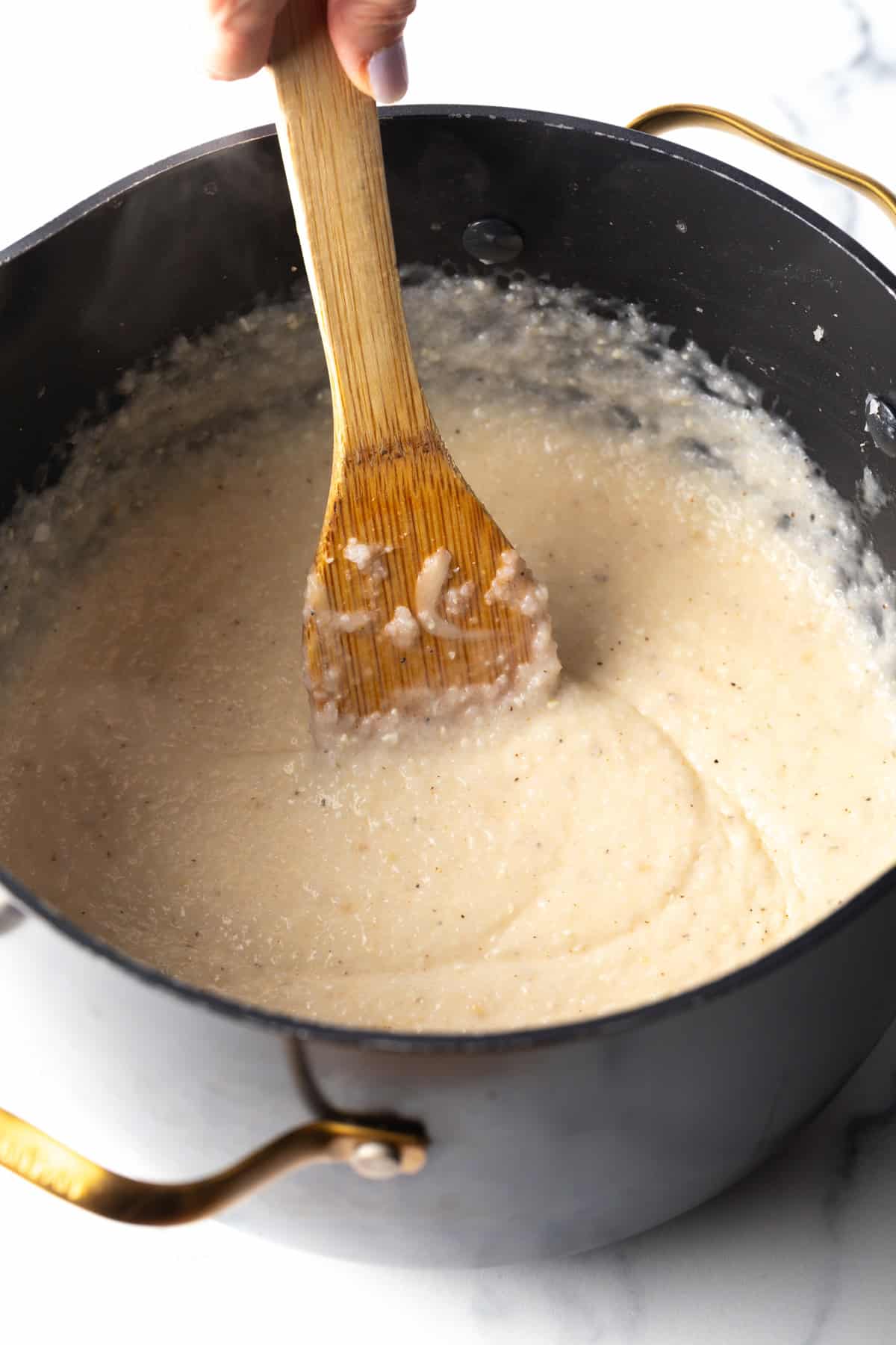 Wooden spatula stirring cheesy grits in a large black cooking pot.