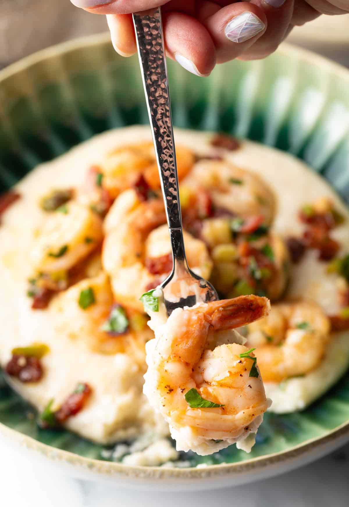 Green bowl filled with cheesy grits and topped with Cajun shrimp and chopped bacon. A spoon with shrimp is being held to camera.