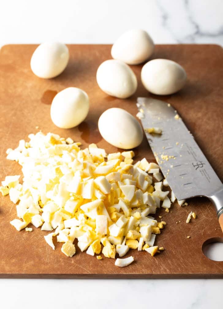 Cutting board with several hard boiled eggs, a pile of chopped hard boiled egg, and a kitchen knife.