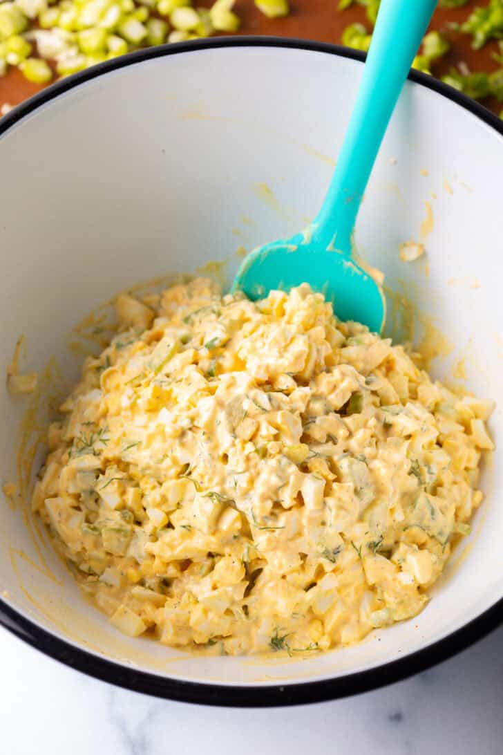 Combining the egg salad mixture in a white bowl with a blue spatula.