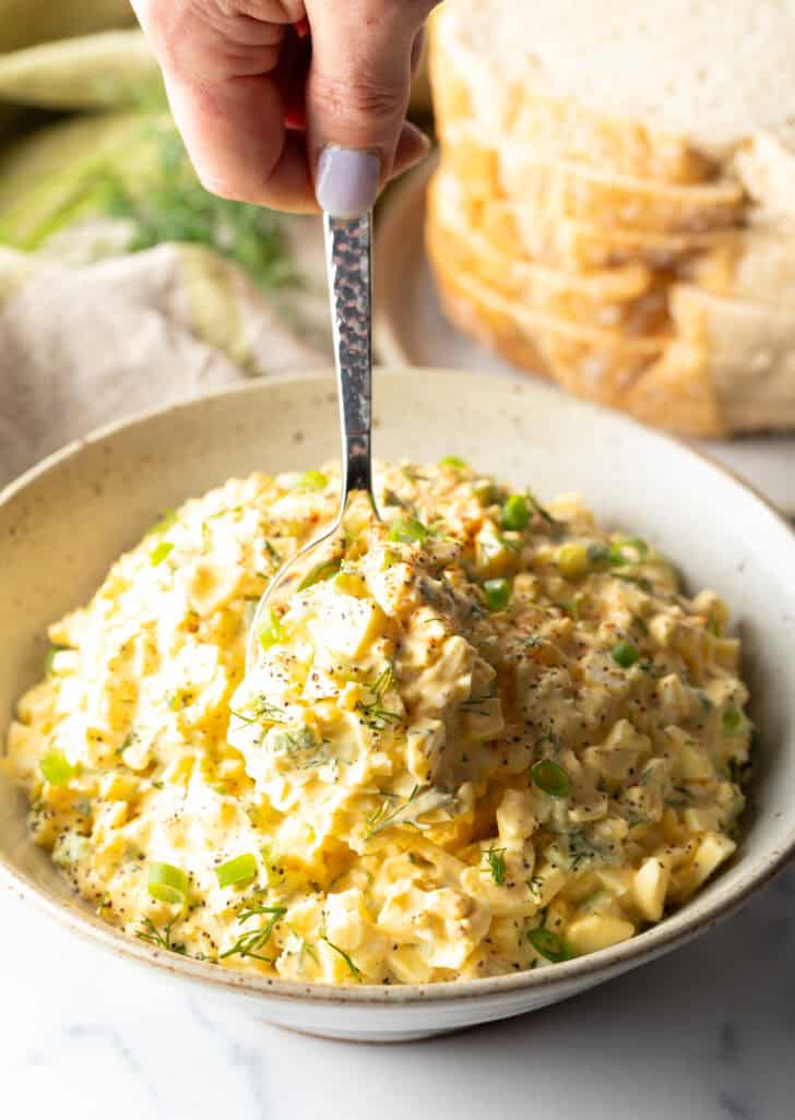 White bowl with egg salad and metal spoon.