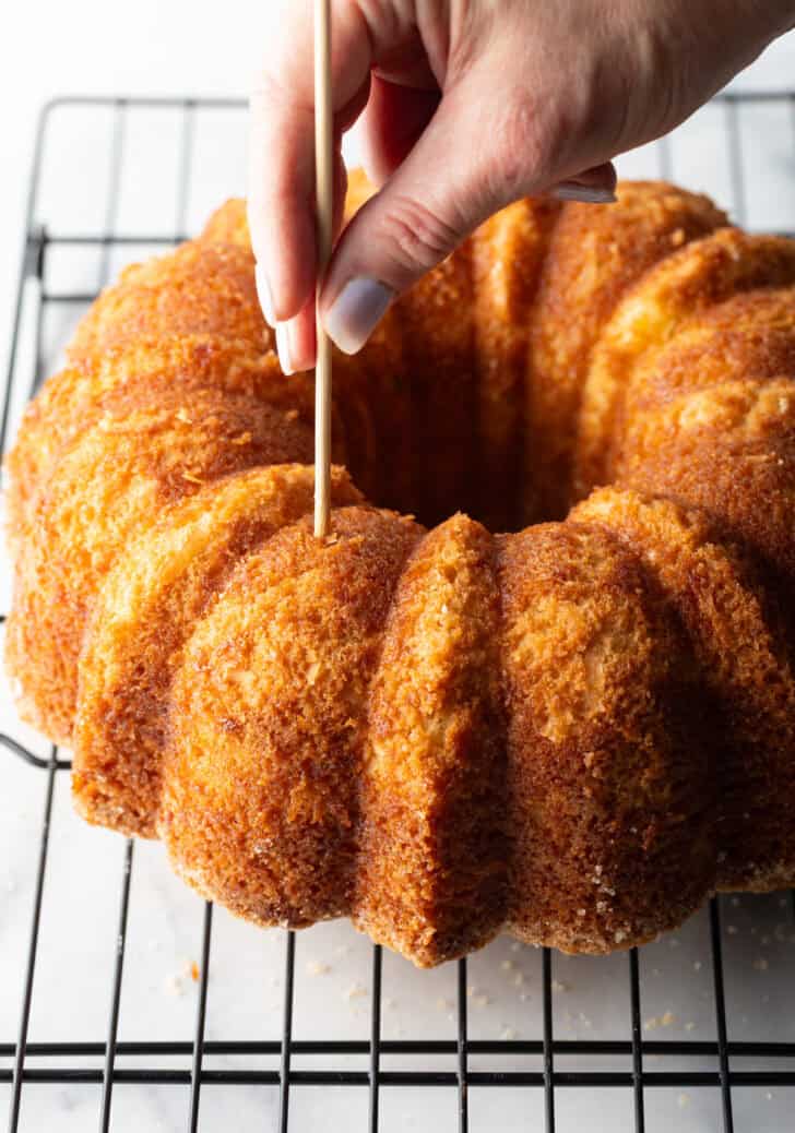 Hand poking holes in a baked bundt cake on a cooling rack.