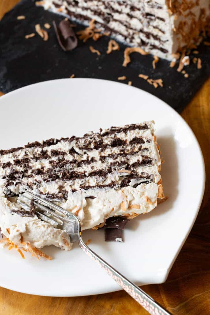 Slice of ice box cake with layers of chocolate graham crackers and Kahlua whipped cream, topped with toasted coconut and chocolate shavings on a white plate. A fork is taking a bite from the bottom of the slice.