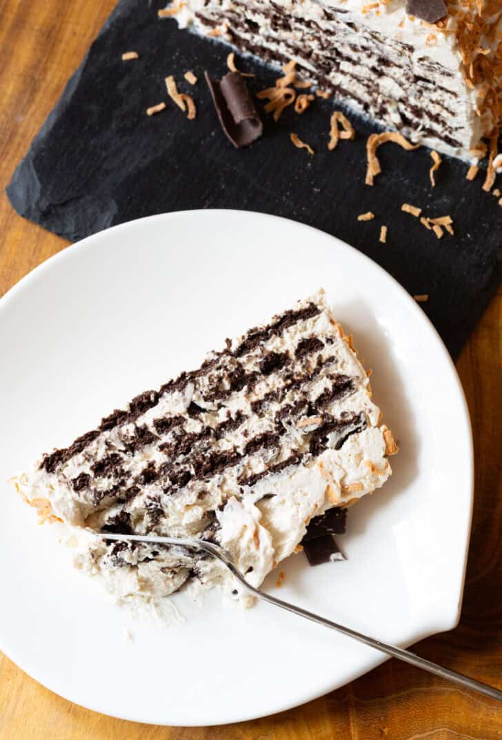 Slice of ice box cake with layers of chocolate graham crackers and Kahlua whipped cream, topped with toasted coconut and chocolate shavings on a white plate. A fork is taking a bite from the bottom of the slice.