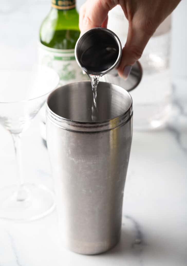 Hand pouring liquid into a metal cocktail shaker from a metal jig.