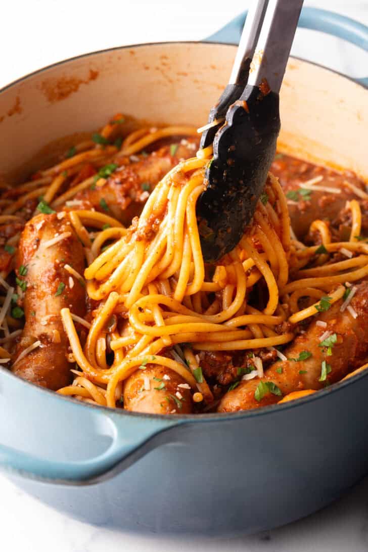Tongs taking a serving of pasta from one pot of spaghetti, sausage, and tomato sauce.