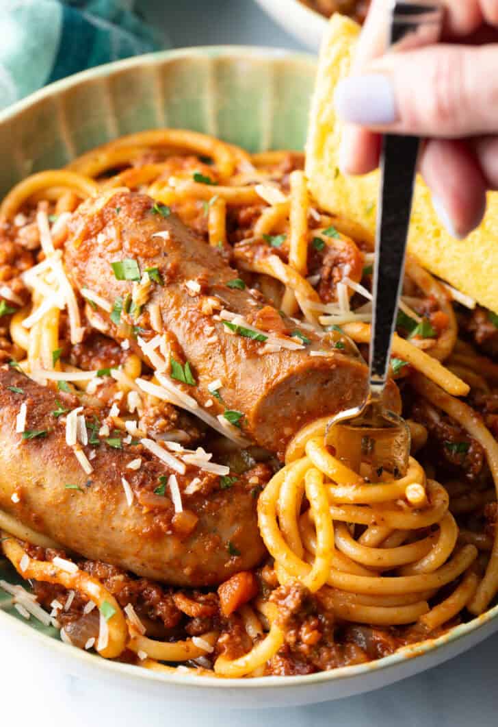 Hand swirling pasta onto a fork from a green bowl of spaghetti and sausage pasta.