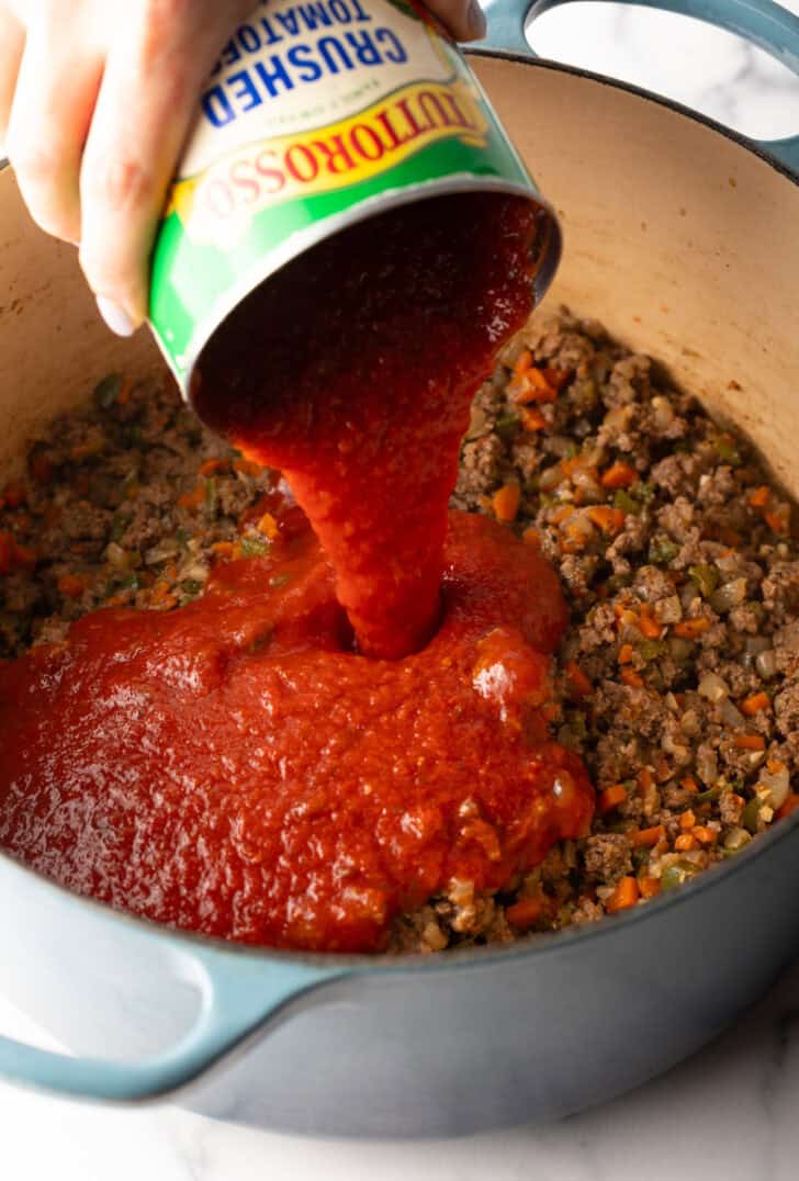 Adding canned tomato sauce to a pot with browned meat and veggies.