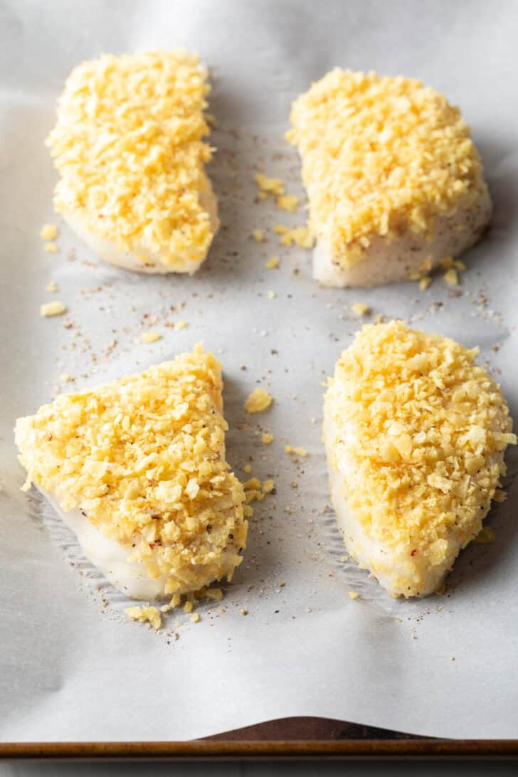 4 baked fish filets coated in crushed potato chips on a baking sheet with parchment paper.