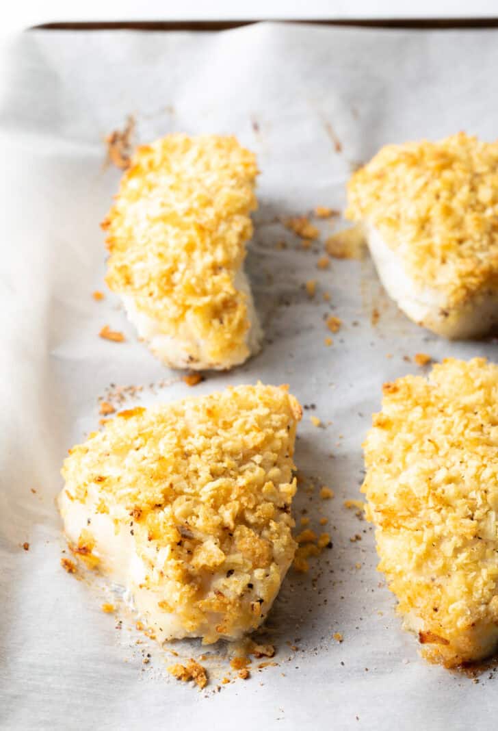 4 baked fish filets coated in crushed potato chips on a baking sheet with parchment paper.