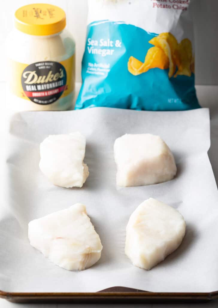 4 pieces of raw sea bass on white parchment, in the background is a bag of salt and vinegar chips and a jar of mayonnaise.
