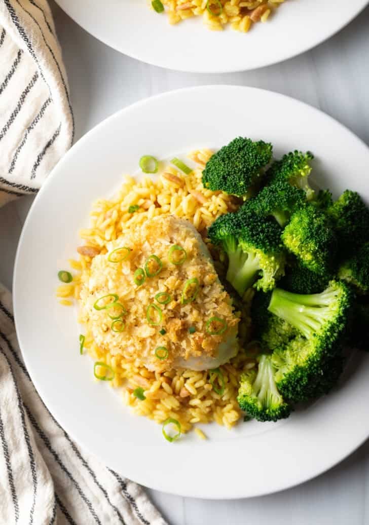 Top down potato chip crusted and baked bass on a plate with yellow rice and cooked broccoli.