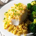 Potato chip crusted and baked bass on a plate with yellow rice and cooked broccoli.