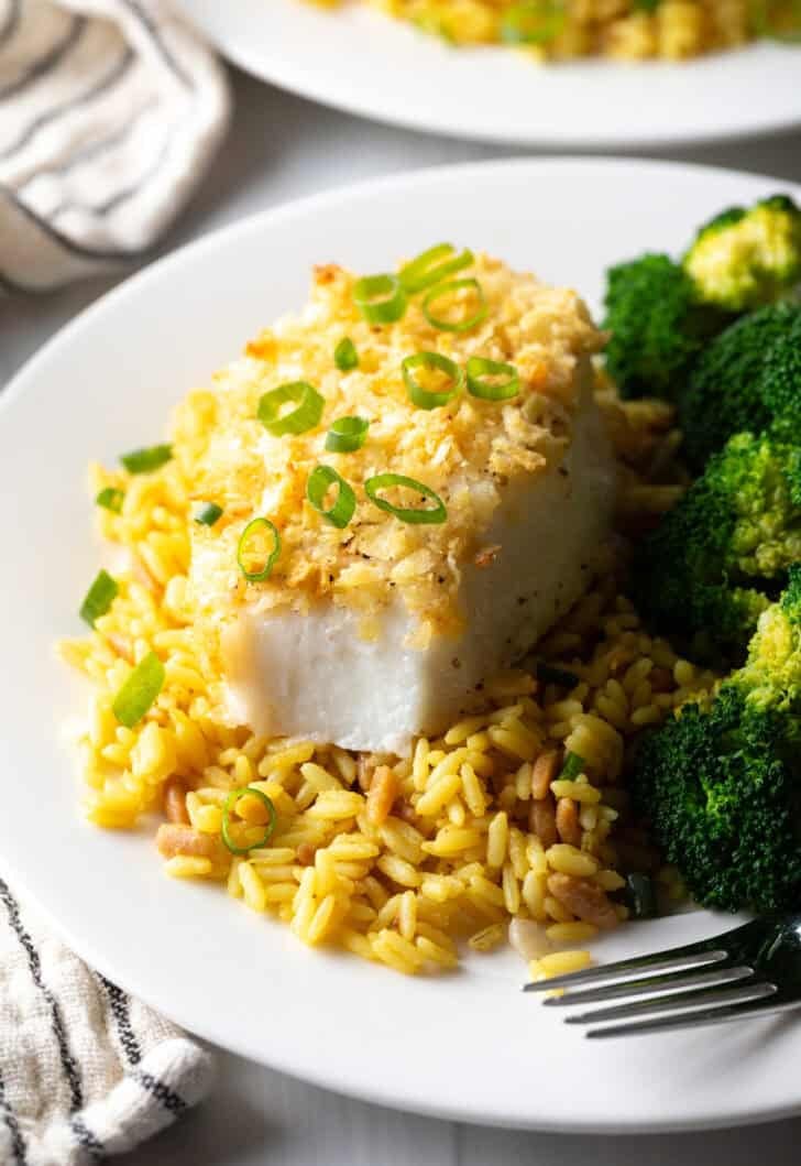 Potato chip crusted and baked bass on a plate with yellow rice and cooked broccoli.