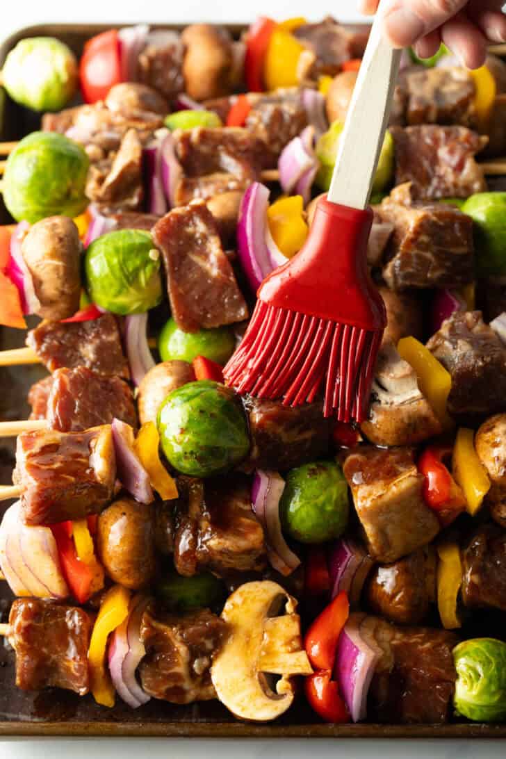 A red silicone brush basting a dozen beef and veggie kebabs that are side-by-side.