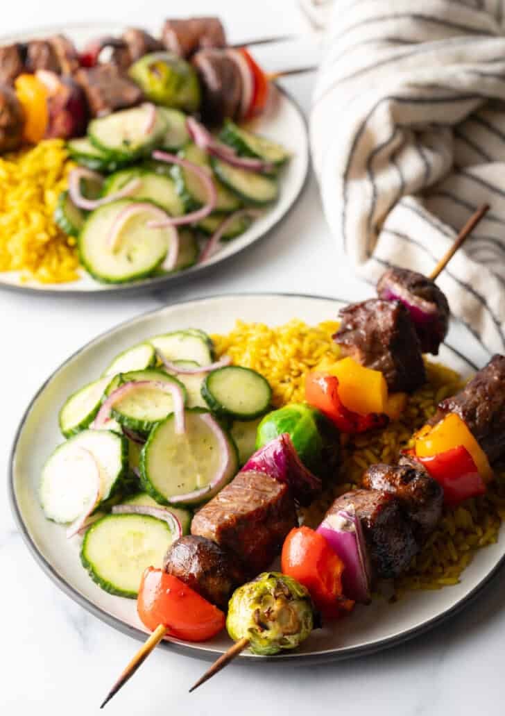 White plate with two veggie and steak beef kabobs, next to a bed of yellow rice and cucumber salad.