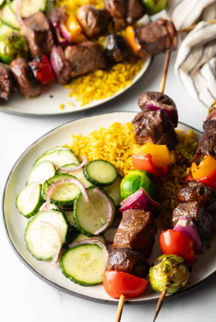White plate with two veggie and steak beef kabobs, next to a bed of yellow rice and cucumber salad.