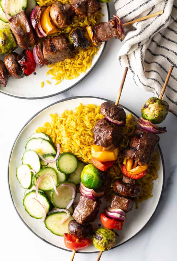 White plate with two veggie and steak beef kabobs, next to a bed of yellow rice and cucumber salad.