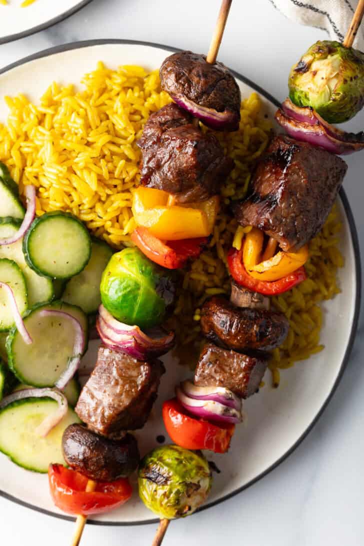 White plate with two veggie and steak beef kabobs, next to a bed of yellow rice and cucumber salad.