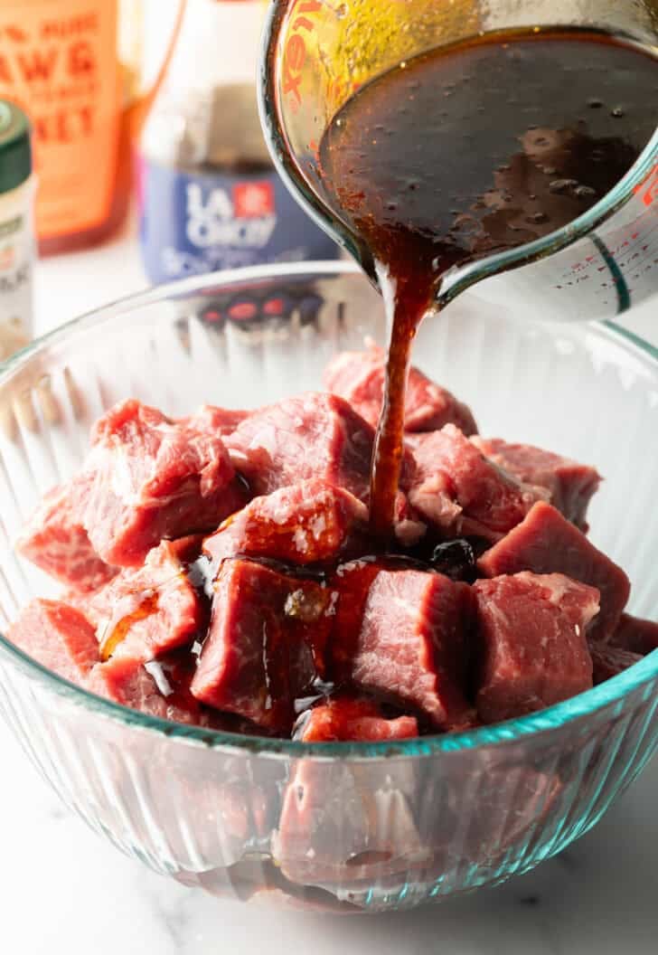 Pouring marinade from glass measuring cup over a glass bowl filled with beef cubes.