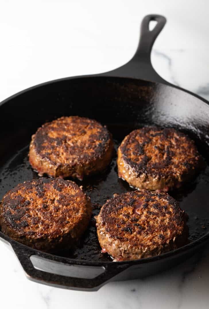 Four cooked hamburgers in a cast iron skillet.