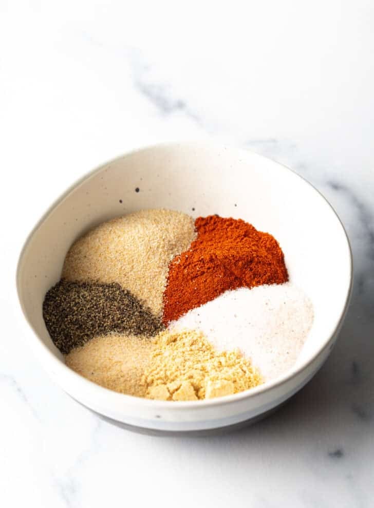 Separate piles of smoked paprika, salt, onion powder, black pepper, garlic powder, ground mustard and a bit of cayenne in a white bowl.
