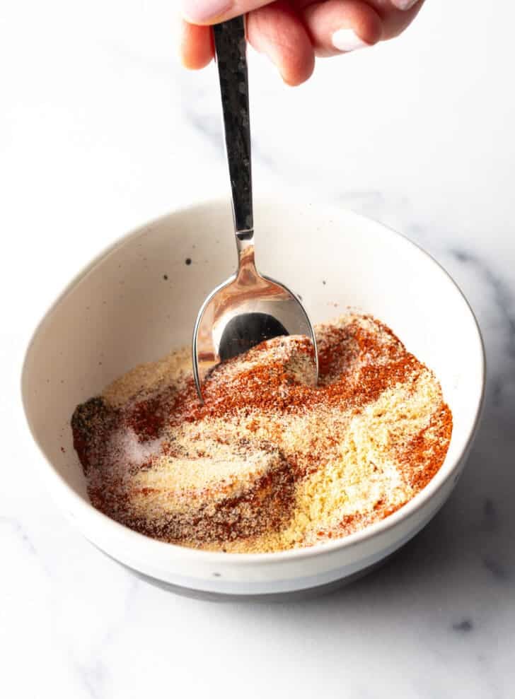 Hand using a spoon to stir together spices in a white bowl.