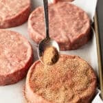 Sprinkling burger seasoning onto a raw hamburger patty with a metal spoon.