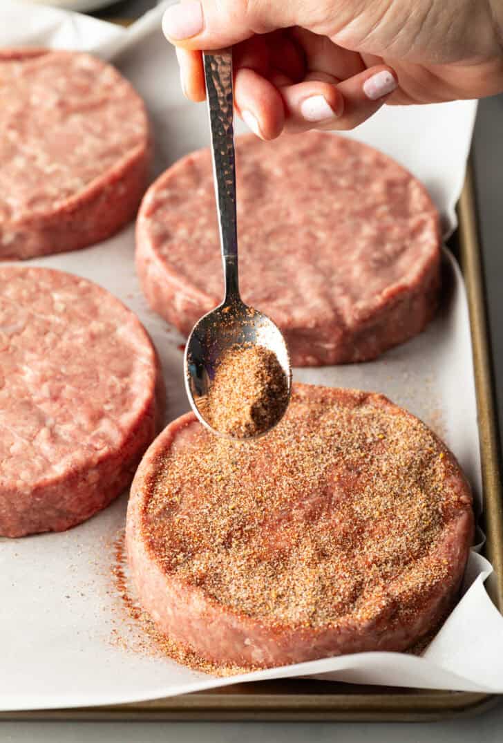 Sprinkling burger seasoning onto a raw hamburger patty with a metal spoon.