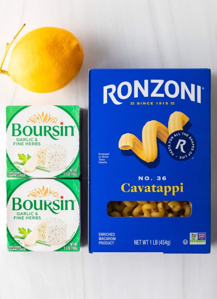 Top down view box of cavatappi pasta, two boxes of Boursin cheese, and a whole lemon.