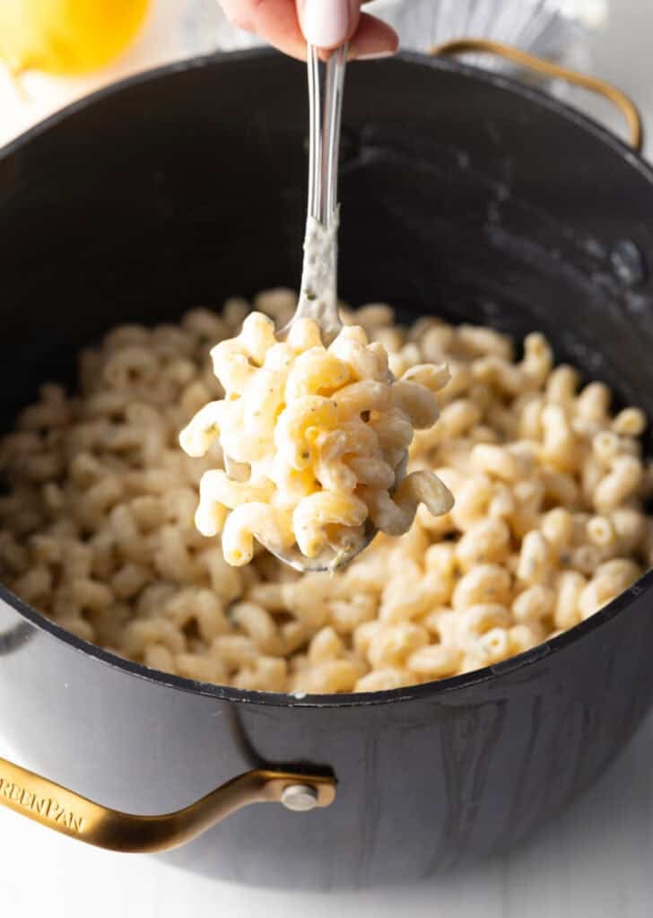 Hand holding a spoonful of cheesy pasta to camera, hovering over a black pot of noodles.
