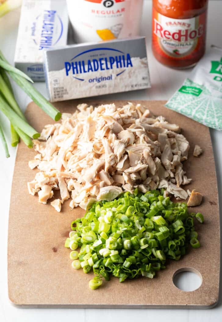 Ingredients you need on a cutting board: Package of cream cheese, cooked shredded chicken, bottle of Franks buffalo sauce, and chopped green onions.