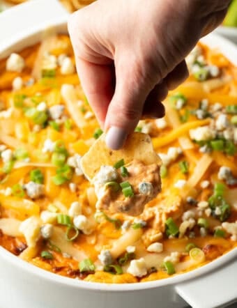 Hand using a tortilla chip to scoop chicken dip from a large white round dish.