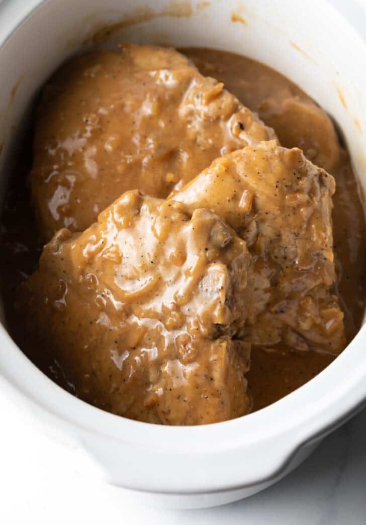 Three large bone-in pork chops in a white crock pot with onion gravy.