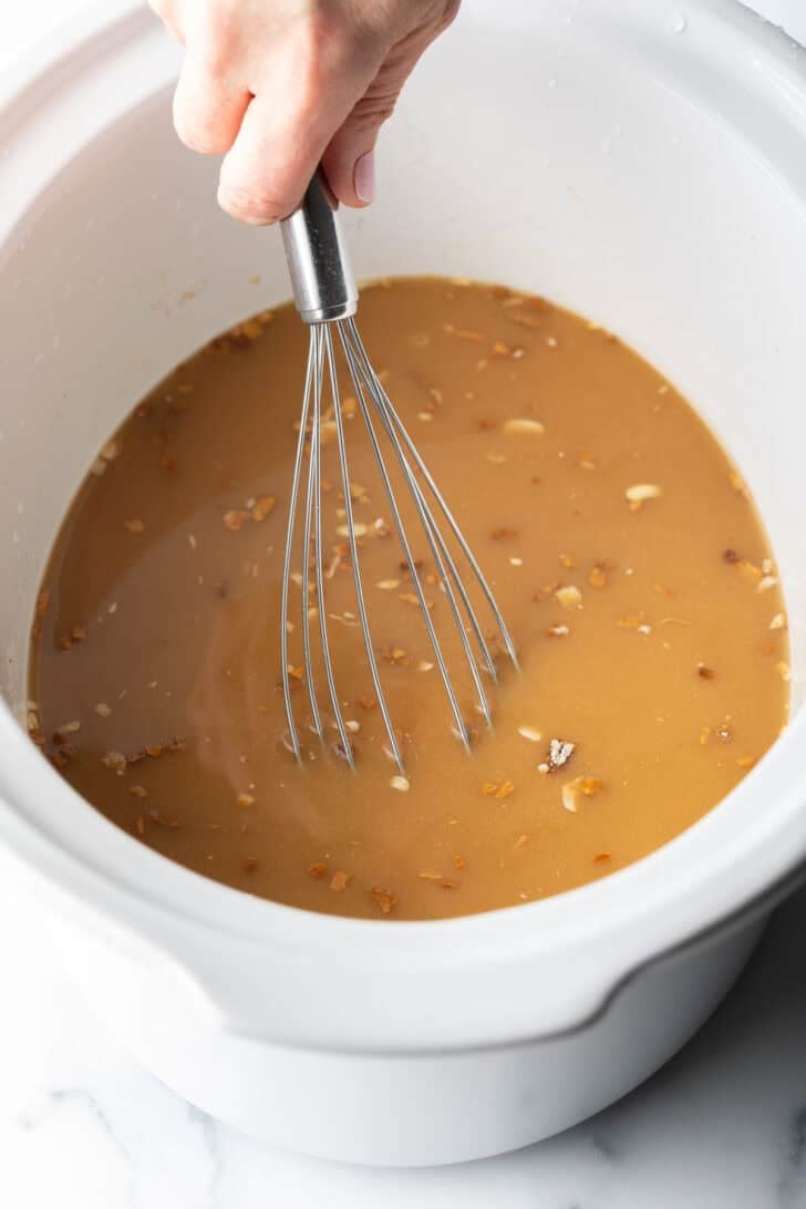 Hand with whisk, whisking brown onion gravy in a white slow cooker.