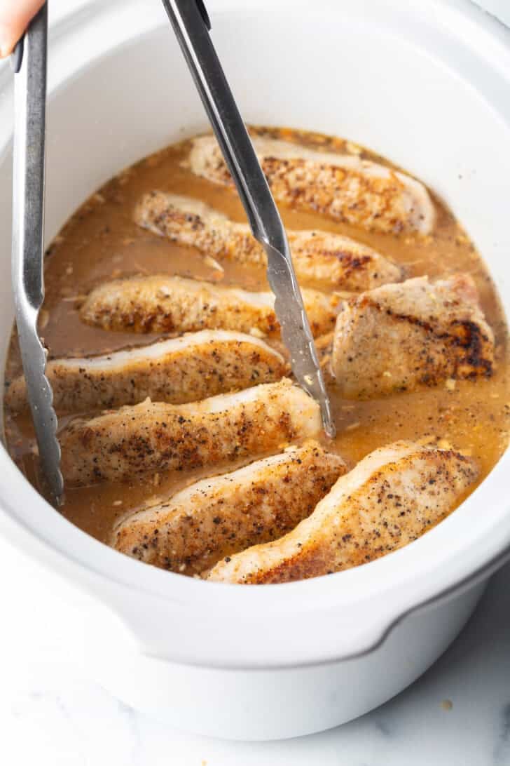 Tongs grabbing a pork chop from a white slow cooker filled with chops and onion gravy.