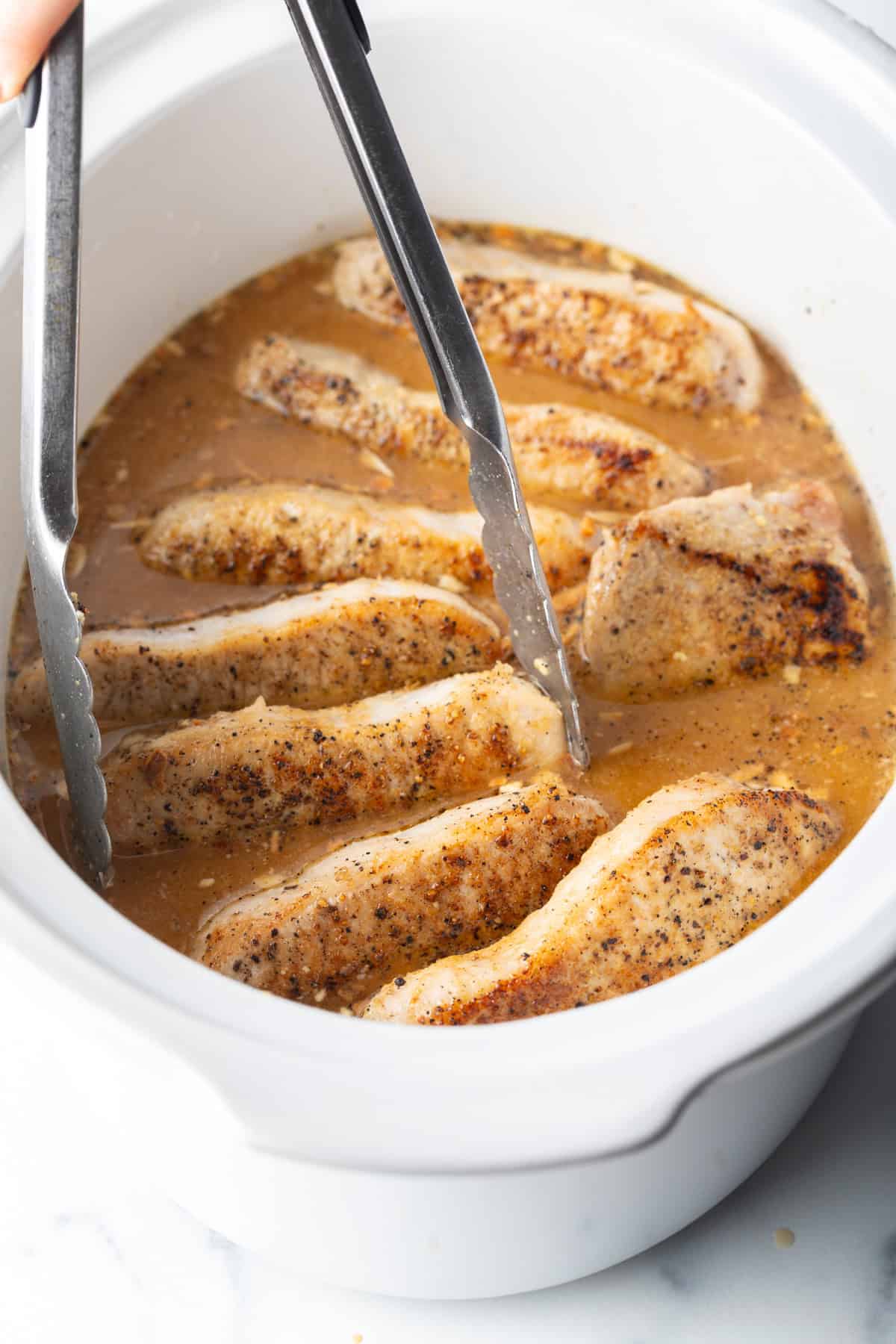 Tongs grabbing a pork chop from a white slow cooker filled with chops and onion gravy.