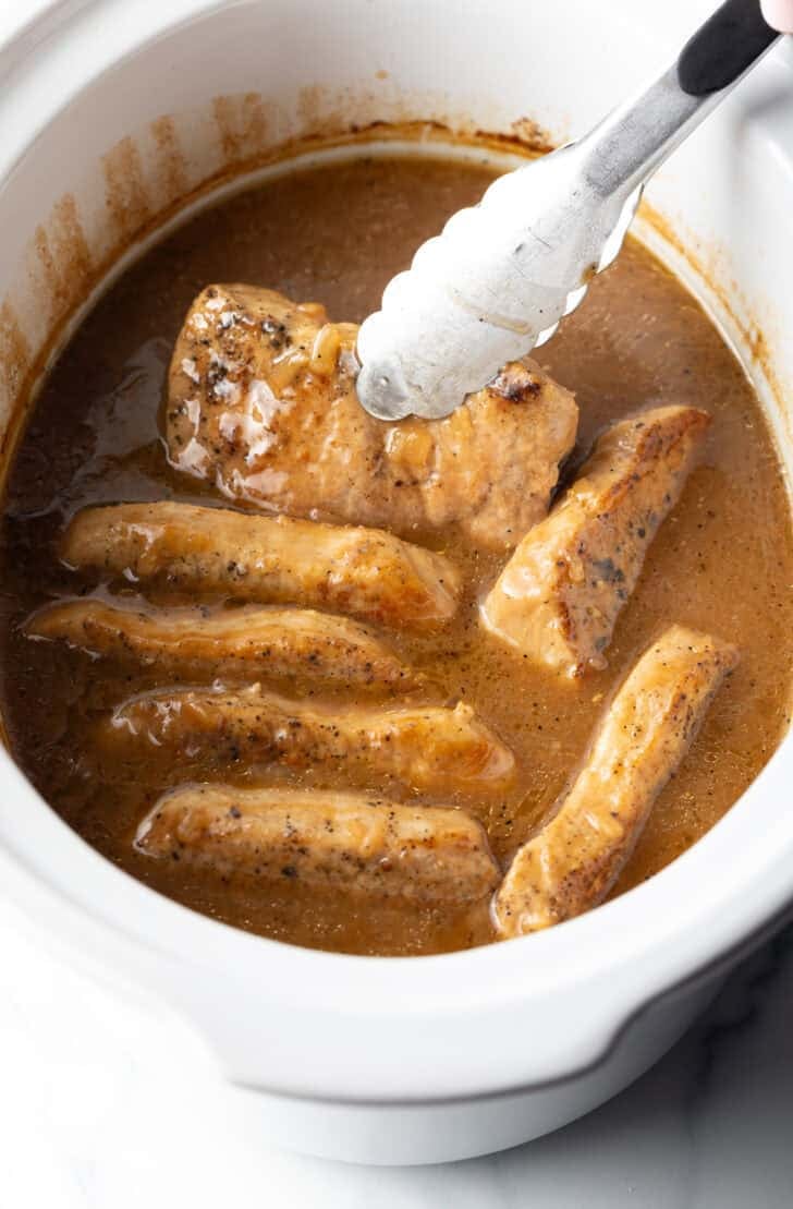 Tongs grabbing a pork chop from a white slow cooker filled with chops and onion gravy.