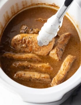 Tongs grabbing a pork chop from a white slow cooker filled with chops and onion gravy.