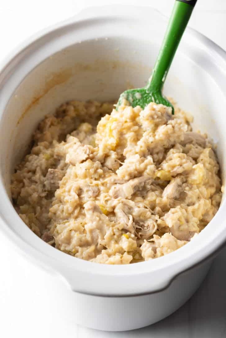 Cooked chicken and rice in a white crockpot, with a green spatula stirring and showing a scoop to camera.